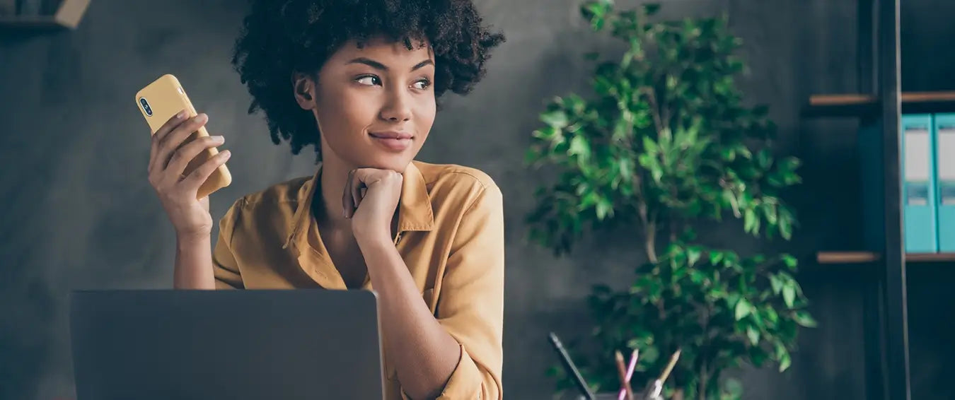 woman holding a cellphone in front of her laptop, looking away with a smile