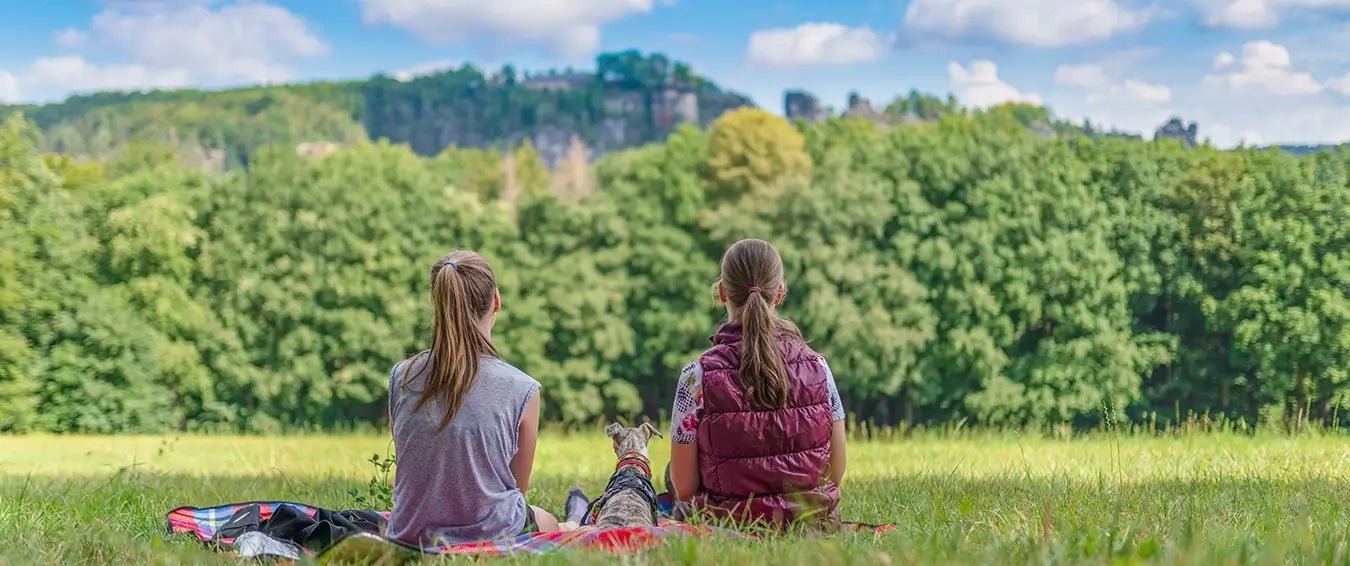 two young woman sitting on a blanket in the nature looking at the green hills