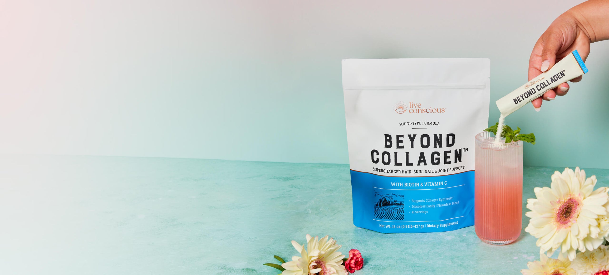 A stand-up pouch of Live Conscious Beyond Collagen powder stands on a light aqua surface beside a pink drink with mint and a hand holding a collagen stick packet above it. White and blue flowers are placed around the setup.